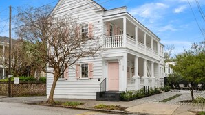 A white colonial-style house with a front porch, a brick wall, and a tree in front. - Designer Chic 6BR Full Downtown Charleston Duplex (Charleston)