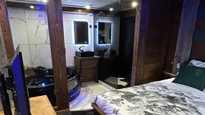 Une chambre à coucher avec un lit en bois, un petit bureau avec un ordinateur, un miroir et une salle de bain avec une douche et des toilettes.