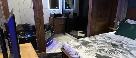 A bedroom with a wooden bed, a small desk with a computer, a mirror, and a bathroom with a shower and toilet.