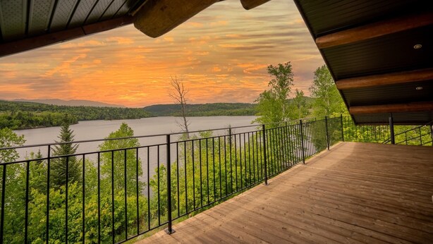 A wooden deck with a metal railing overlooking a lake and forested hills during sunset.