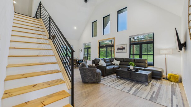 A spacious living area with a wooden staircase, a sectional sofa, a coffee table, and large windows offering views of the outdoors.