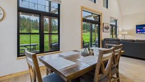 A dining area with a large table and chairs, a view of a grassy field through large windows, and a modern living space with a sofa and a TV.