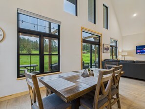 A dining area with a large table and chairs, a view of a grassy field through large windows, and a modern living space with a sofa and a TV. - Buffalo Bungalow - 2 BD Cabin (Custer)