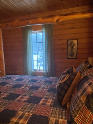A cozy cabin room with a plaid bedspread, a window with curtains, and a framed picture on the wall.