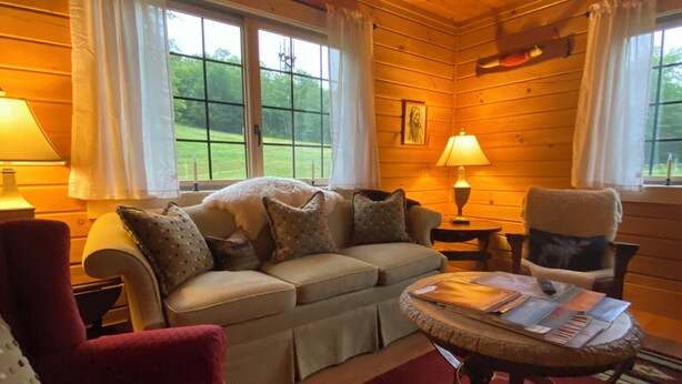Interior acolhedor de uma cabana, com teto de madeira, um sofá, uma poltrona vermelha, uma mesinha com revistas e uma janela com cortinas.