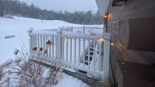 Una terraza cubierta de nieve con barandilla blanca y luces de cuerda, que conduce a una cabaña de madera con base de piedra.