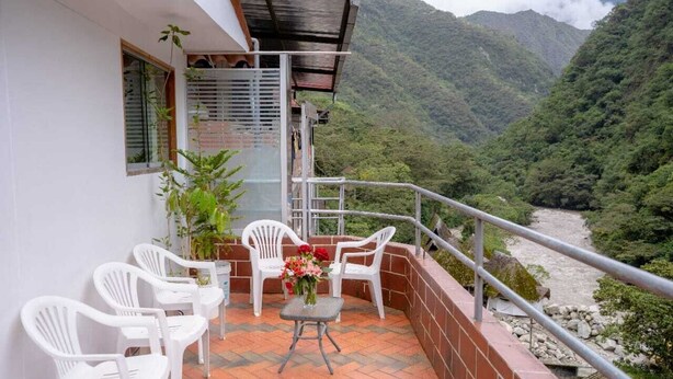 A balcony with white plastic chairs and a table, overlooking a river and mountains. - Hotel Vista Mapi (Machu Picchu)