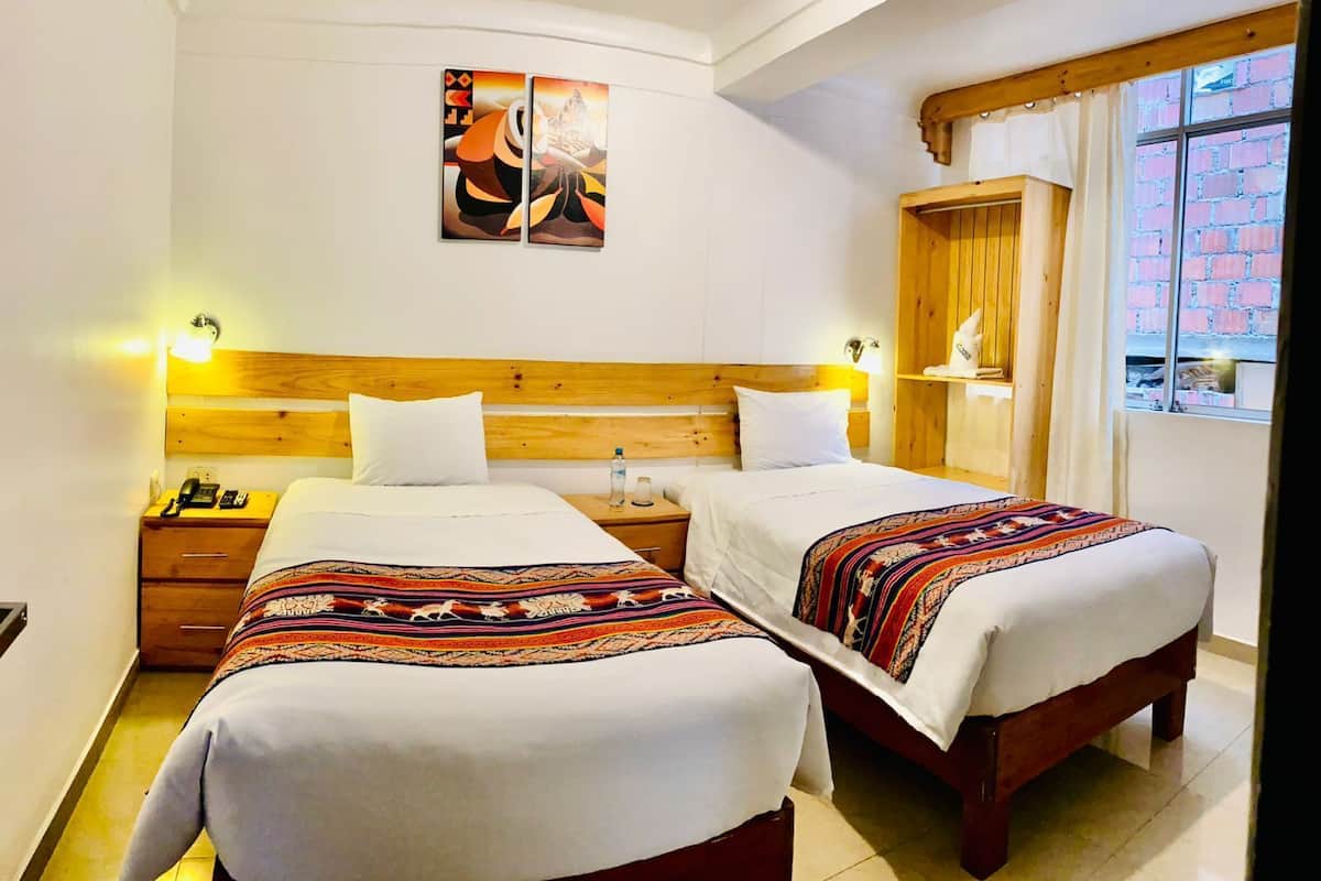 Ein Hotelzimmer mit zwei Betten, Holz-Kopfteilen, einem Bild über den Betten, einem Nachttisch mit Telefon und Lampe sowie einem Fenster mit Blick auf ein Backsteingebäude.