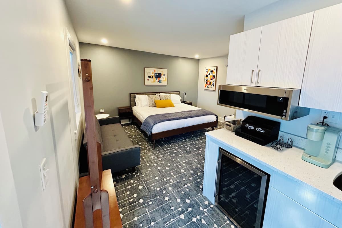 Una habitación de hotel moderna con cama, microondas, cafetera, lavamanos y un pequeño rincón de descanso.