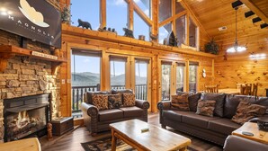 A cozy log cabin interior with a stone fireplace, leather sofas, and a large window overlooking a mountain view.