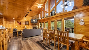 A wooden cabin interior with a fireplace, a television, a dining area with wooden chairs and a table, and a large window overlooking greenery.