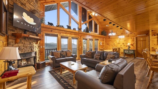 A cozy log cabin interior with a fireplace, leather sofas, wooden tables, and a large TV screen displaying the Magnolia logo.