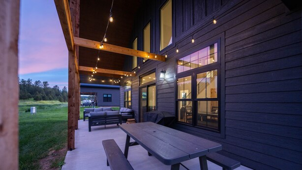 An outdoor patio with a wooden table and benches, string lights, and a view of a house with large windows. - Custer's Retreat - 2 BD Cabin (Custer)