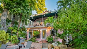 A patio with outdoor furniture and a grill, surrounded by lush greenery and a pink house with a balcony. - 3 Bedroom Home - Perfect Location & Private Pool (Key West)