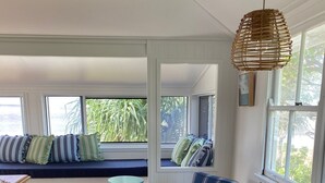 Een kamer met een houten vloer, rieten stoelen, een bank met kussens en grote ramen.