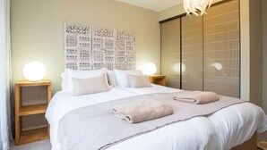 Ein ordentlich bezogenes Bett mit zwei Kissen und einem gefalteten Handtuch, ein Nachttisch aus Holz mit einer runden Lampe und eine dekorative Wandverkleidung.