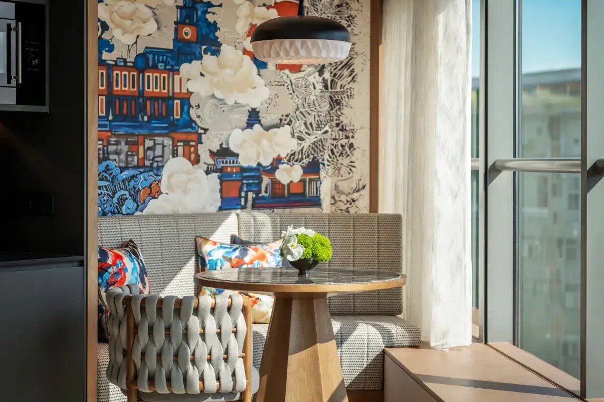 A modern room with a large window, a sofa with patterned cushions, a round wooden table with a vase of flowers, and a wall mural depicting a cityscape.