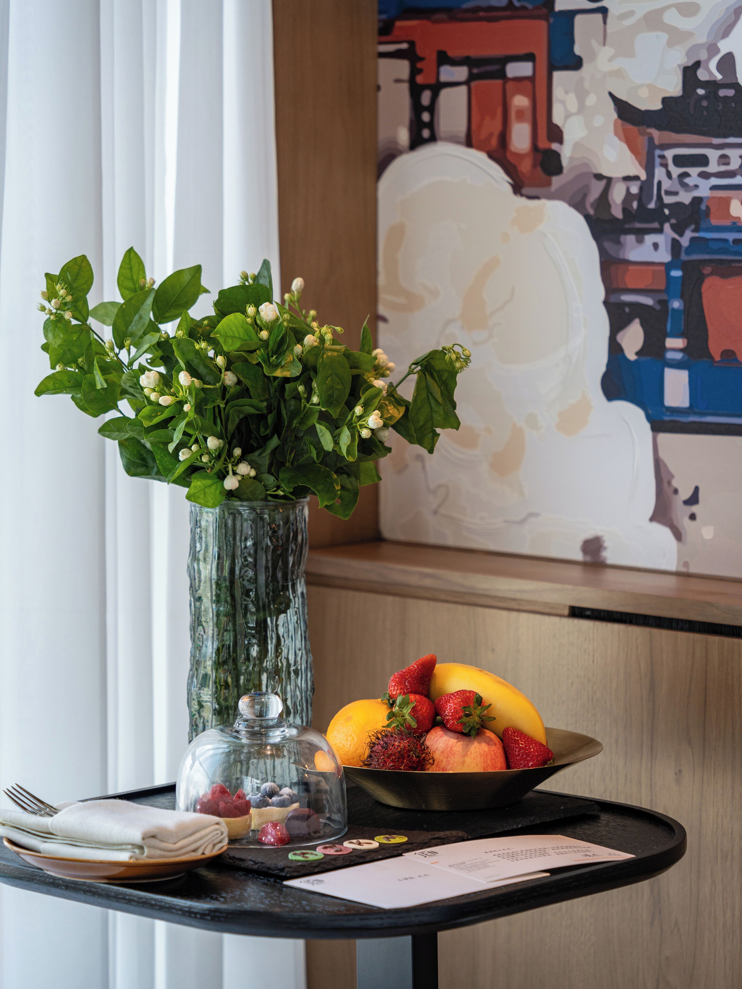 A table with a vase of green leaves, a bowl of fruit, and a glass dome over a decorative item.
