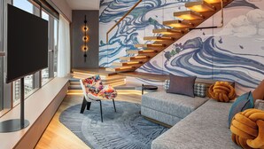 Una sala moderna con un televisor grande, un sillón colorido, un sofá con un cojín original y una escalera con un muro detrás con un diseño ondulado.