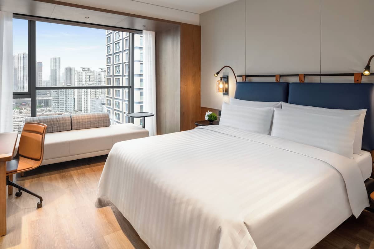 Une chambre d’hôtel moderne dotée d’un grand lit, d’un coin salon avec vue sur la ville et d’une petite table avec une lampe.