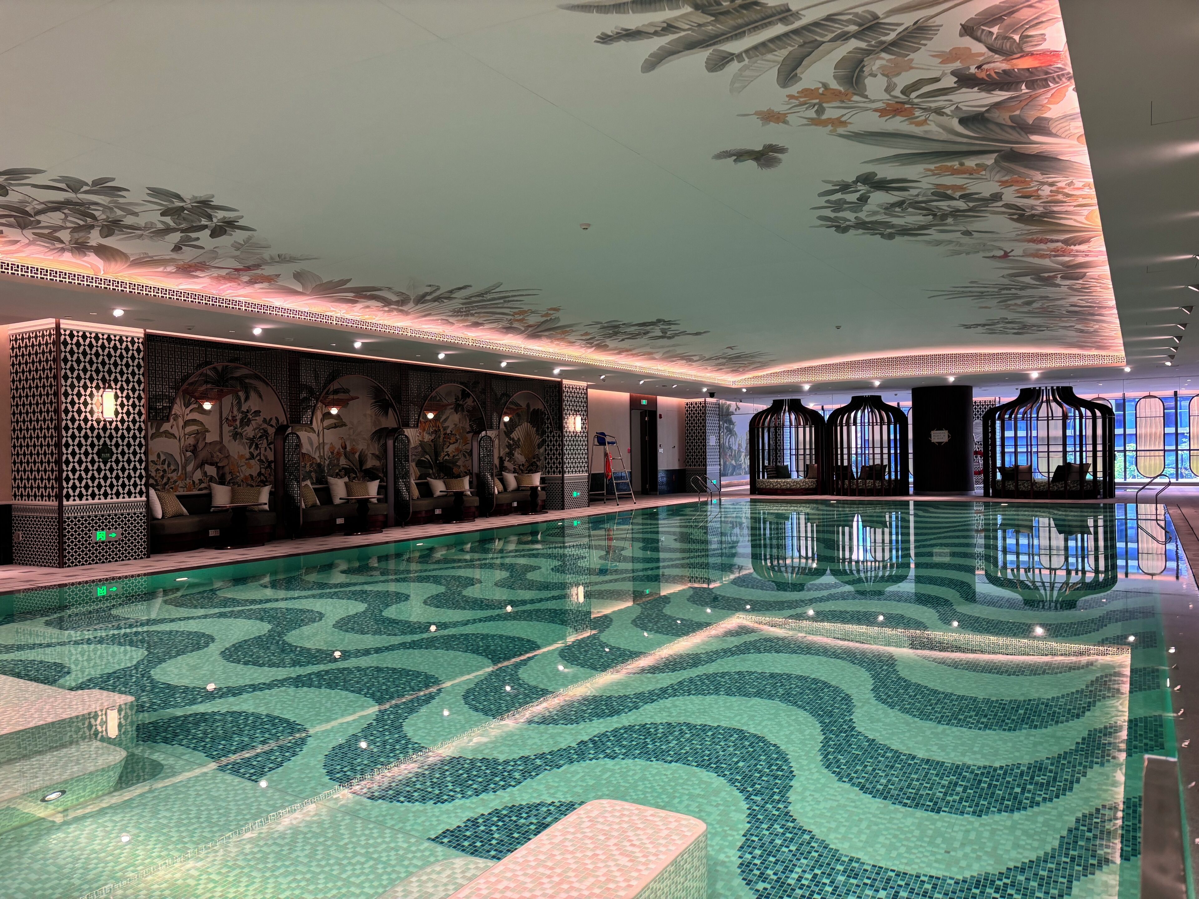 Una amplia piscina interior con un diseño decorativo en el suelo, zonas de estar con cojines y un mural con motivos florales y de pájaros en las paredes.
