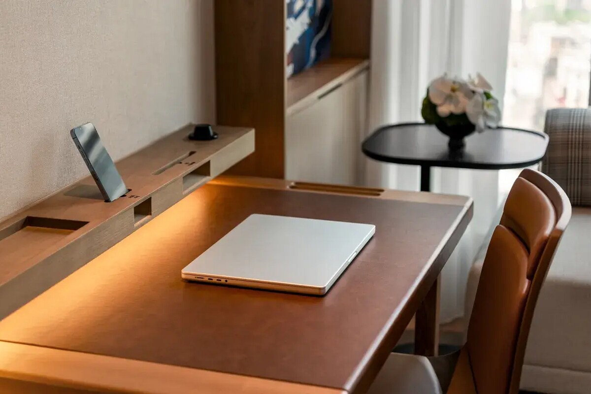A wooden desk with a laptop, a smartphone, and a small speaker. There is a chair and a round table with a vase of flowers in the background.