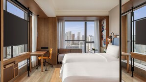 Habitación de hotel con dos camas, un escritorio, una silla, una mesita y un amplio ventanal con vista a la ciudad.
