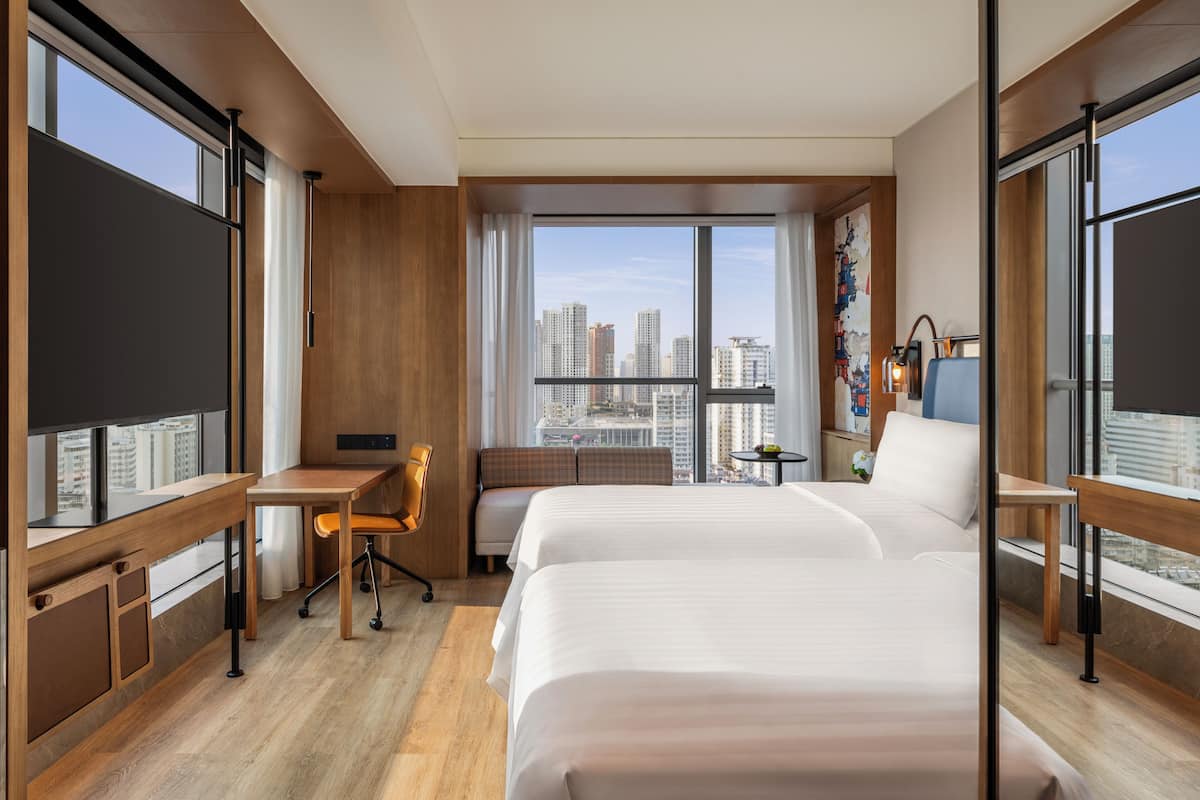 Une chambre d’hôtel avec deux lits, un bureau, une chaise, une petite table et une grande fenêtre donnant sur la ville.