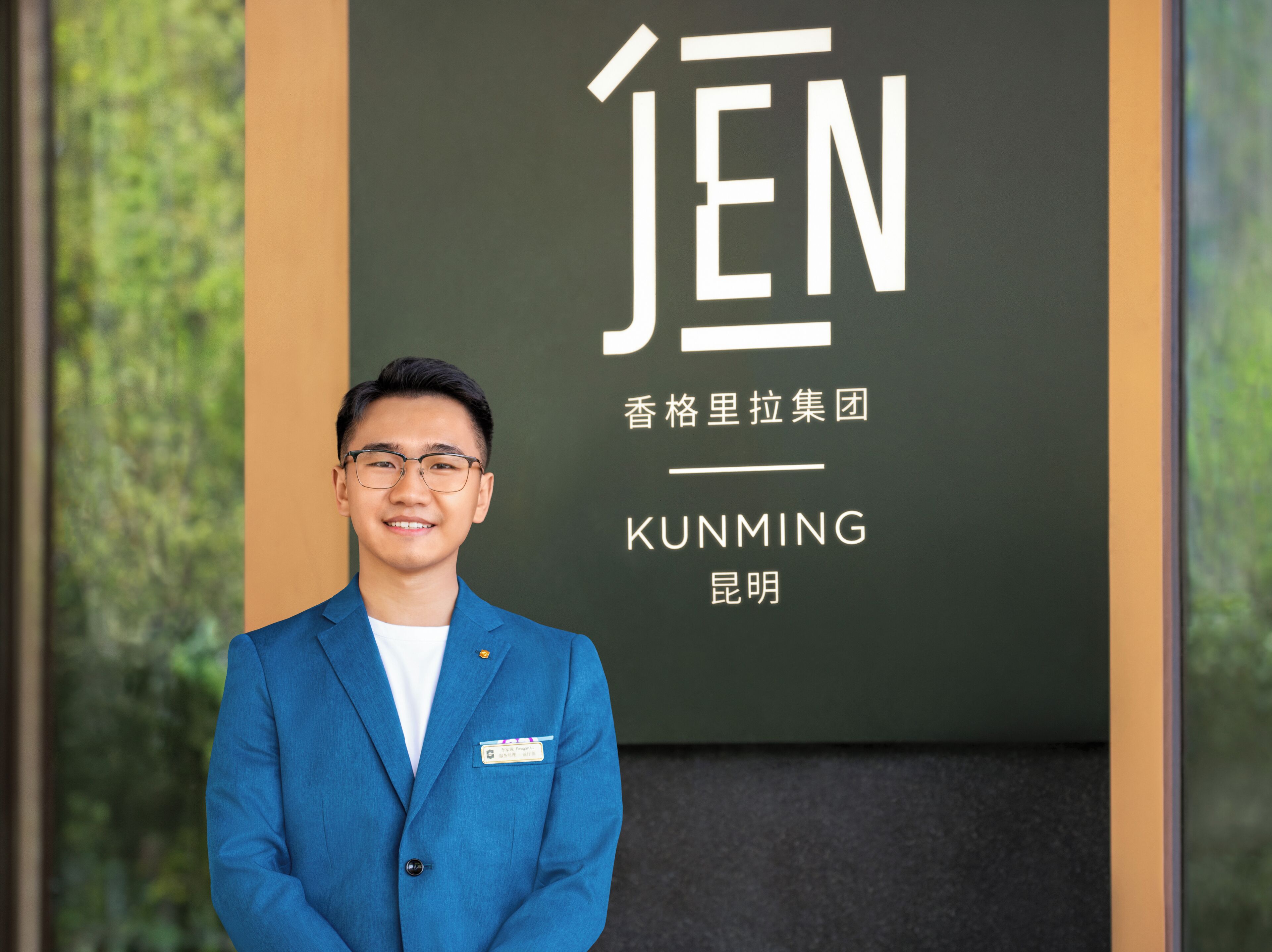 A man in a blue suit standing in front of a sign with the word "JEN" and some Chinese characters.
