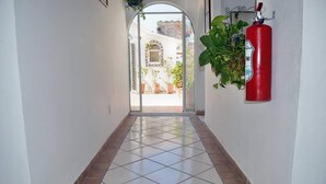 Un couloir carrelé mène à un espace ouvert où se trouvent des plantes en pot et un extincteur fixé au mur.