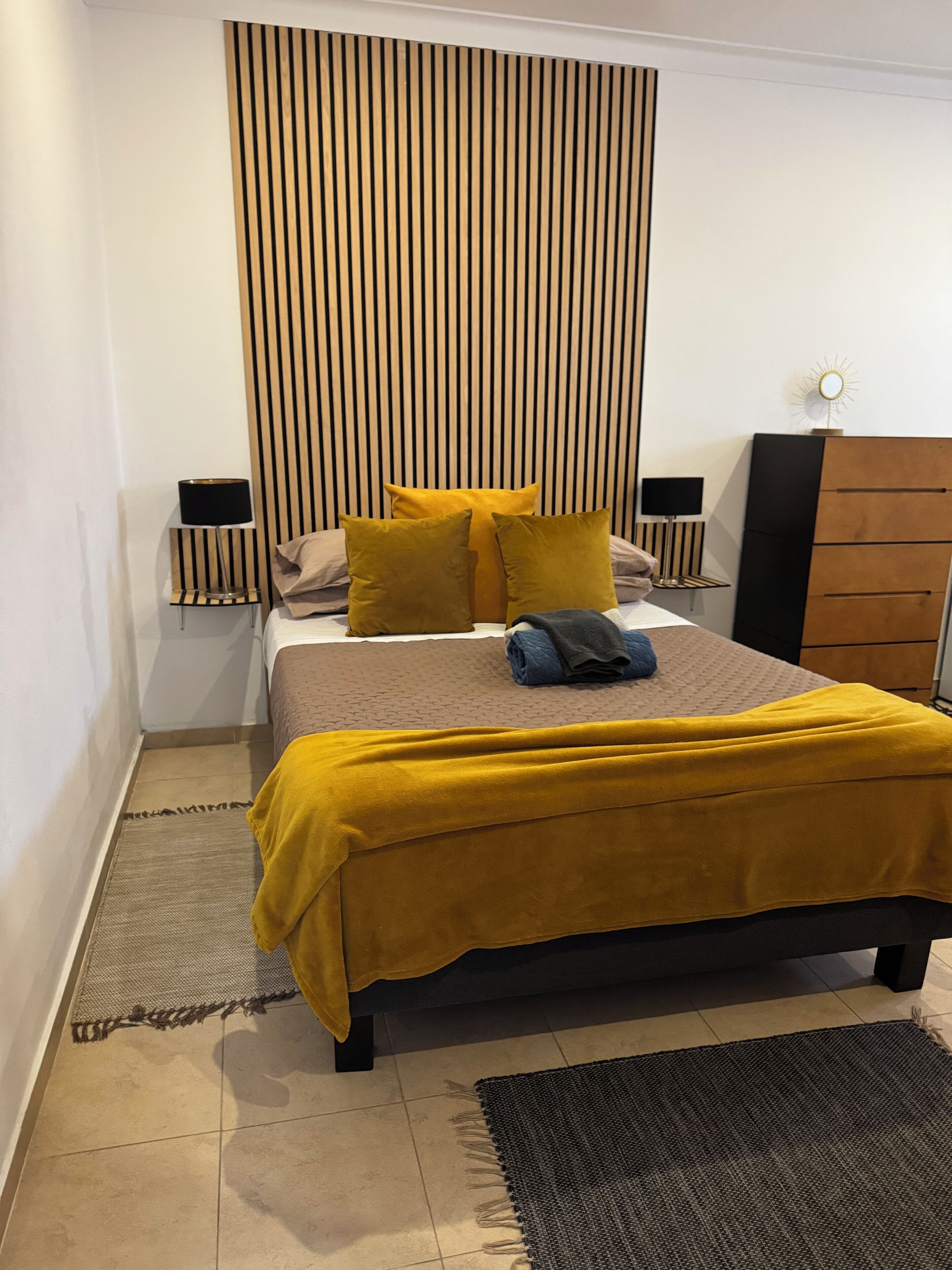 Un dormitorio moderno con cama, cabecera con líneas verticales, mesita de noche, cómoda y una pequeña mesa con lámpara.