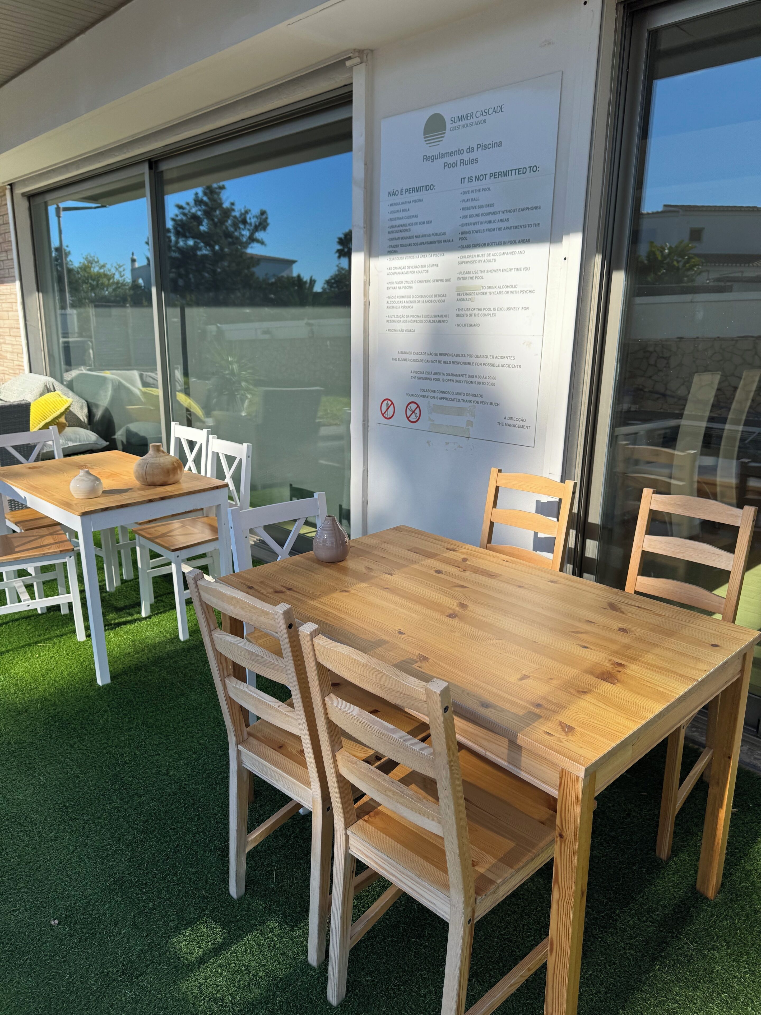 An outdoor seating area with wooden tables and chairs, a glass wall, and a sign with pool rules.