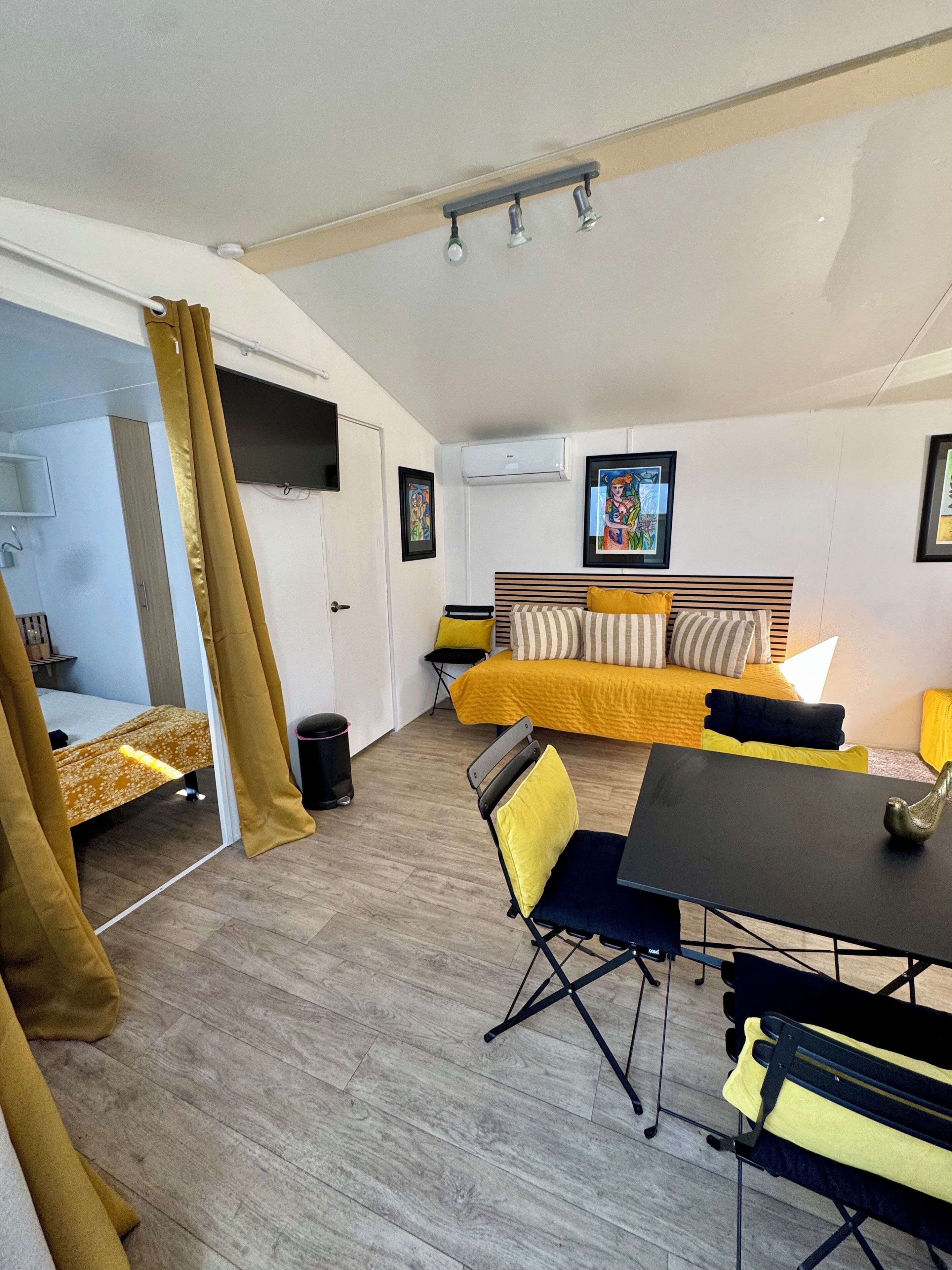 Een compacte kamer met een bed, een slaapbank, een eettafel en een stoel. Er is een raam met gordijnen, een wandairconditioner en een fotolijst.