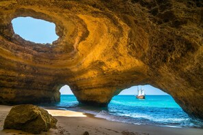 A natural rock formation with a cave-like opening, a sandy beach, and a sailboat in the water.