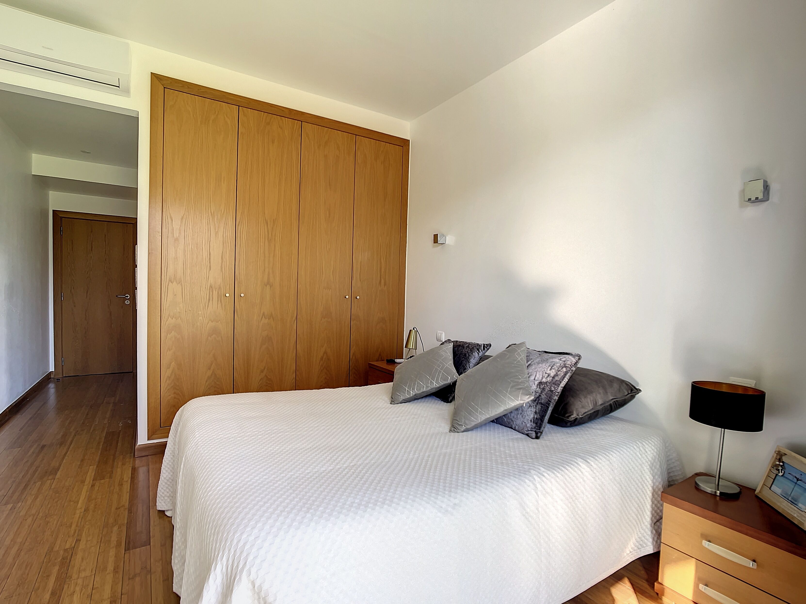 A bedroom with a large wooden wardrobe, a bed with a white bedspread and grey pillows, a bedside table with a lamp, and a small drawer unit.