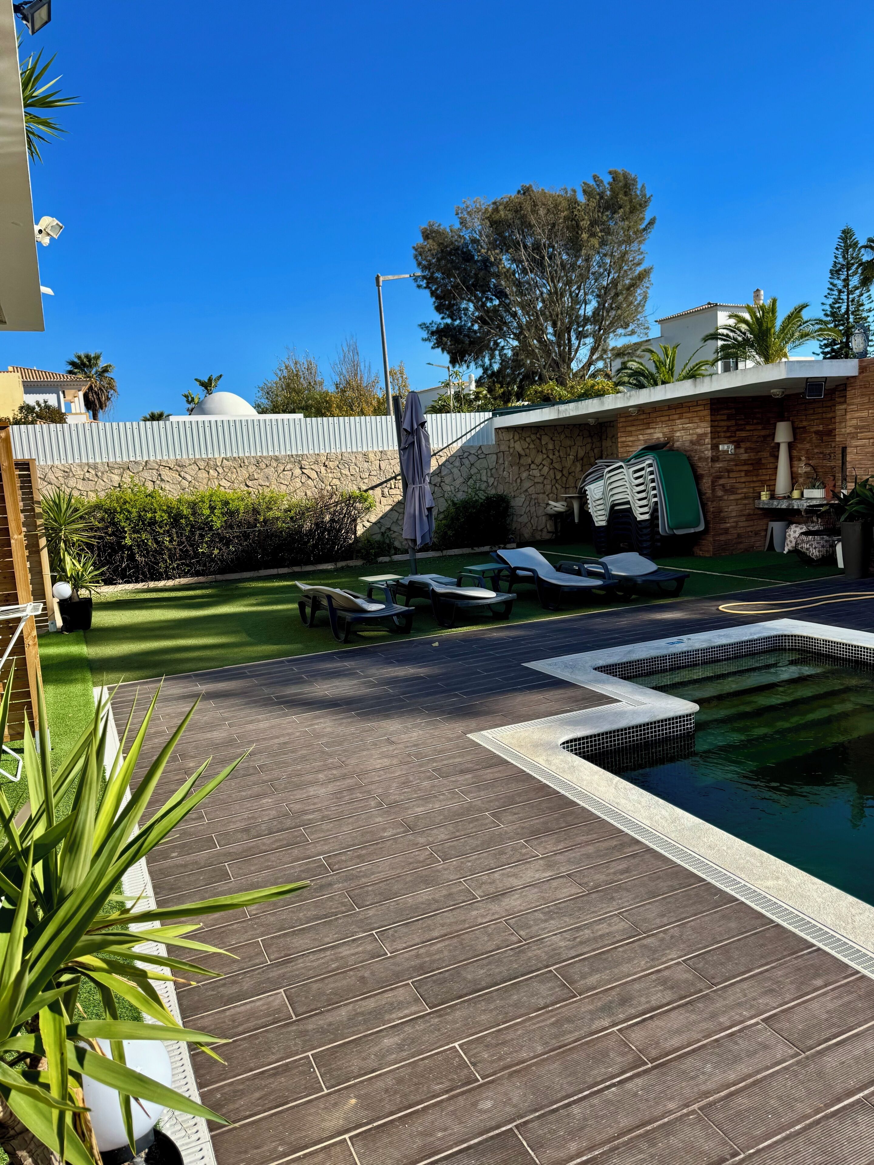 Piscina com espreguiçadeiras, uma parede de pedra e uma área coberta com um candeeiro.
