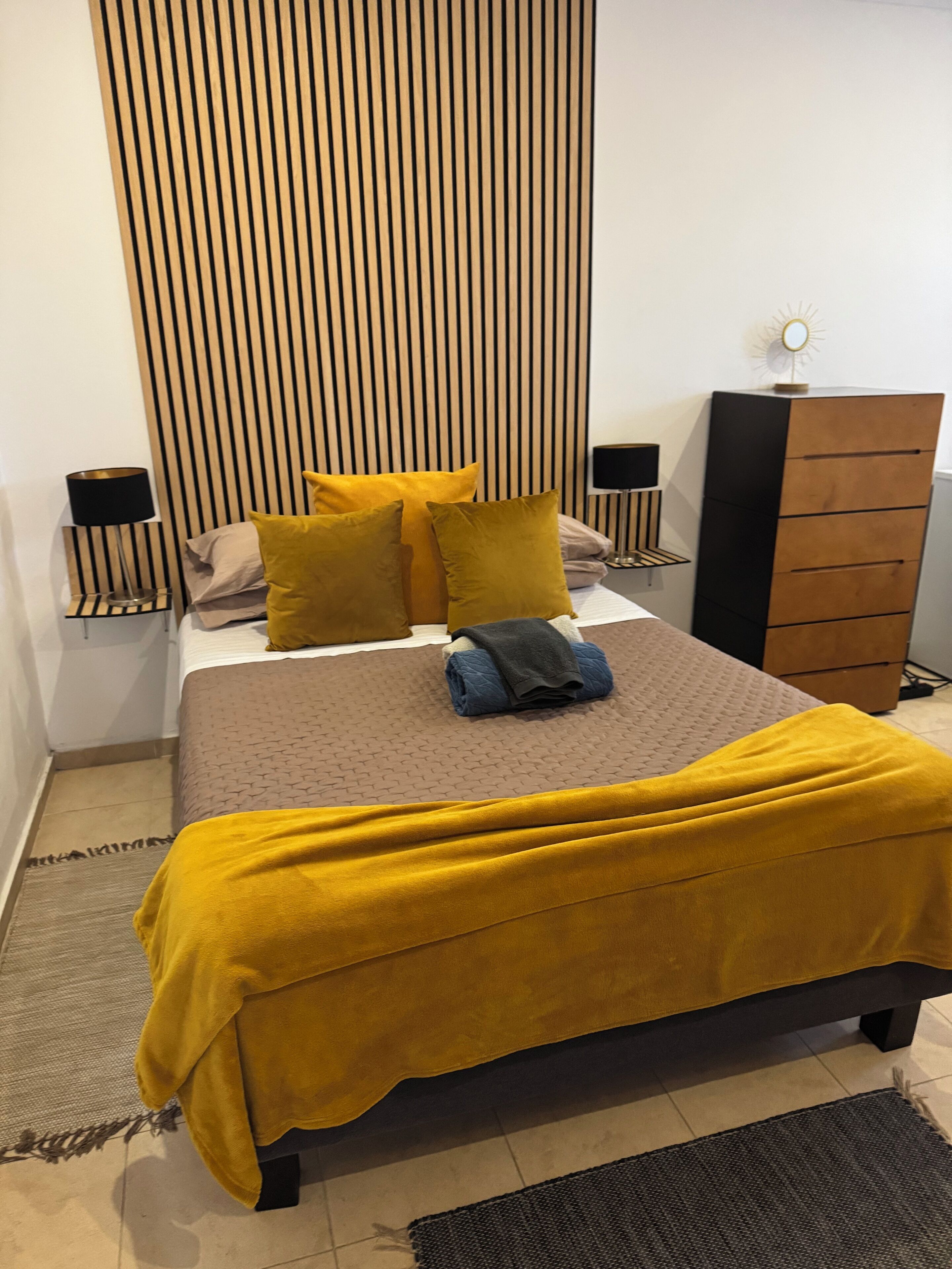 A bedroom with a bed, a nightstand, a dresser, and a wall with vertical lines.
