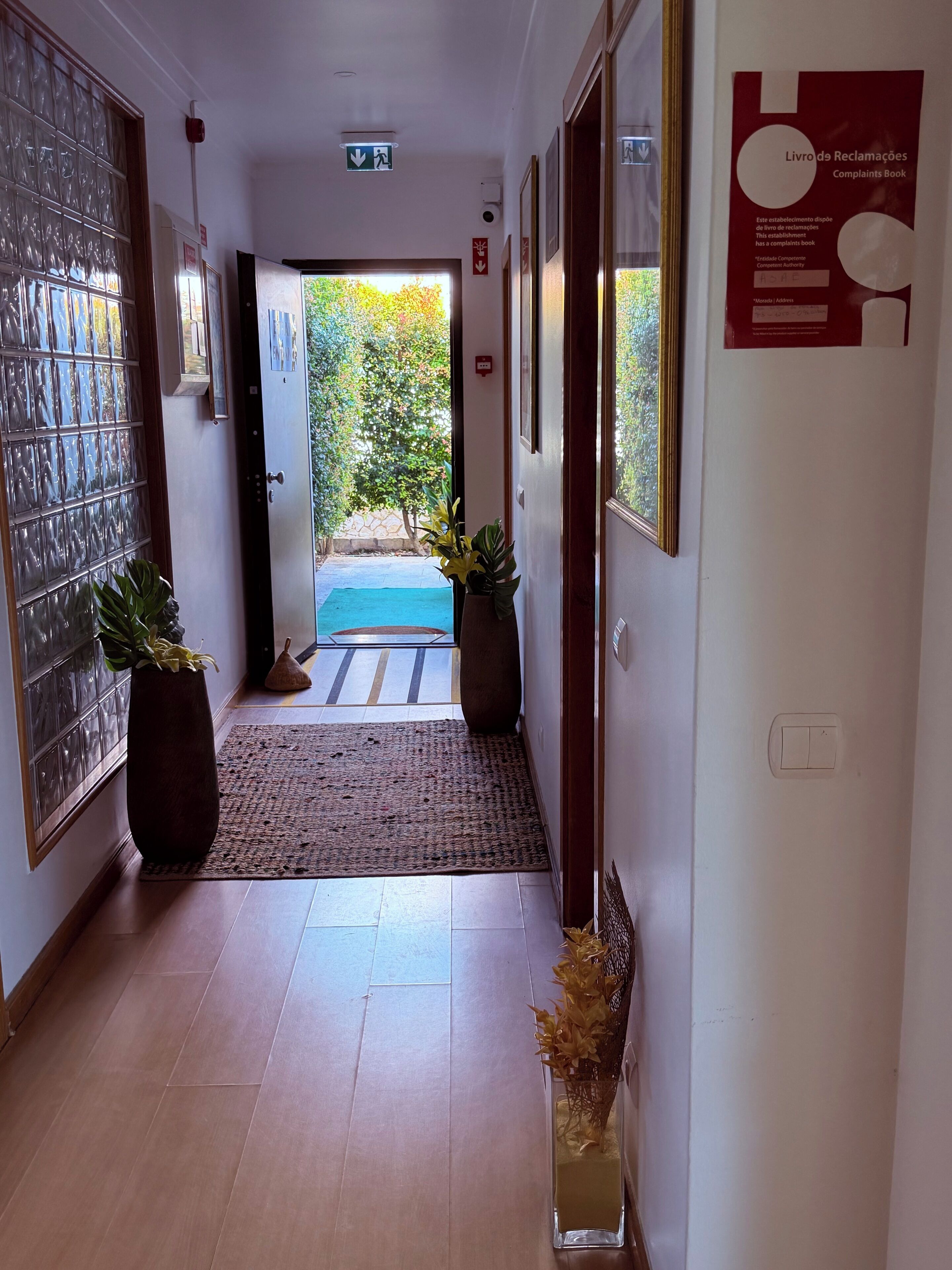 Un pasillo con una puerta de cristal, un libro de reclamaciones y vistas a un jardín.