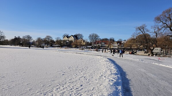 A snow-covered park with a wide open area, a few people walking, and buildings in the background. - Attic Floor With Views Over Fields and sea (Stockholms län)