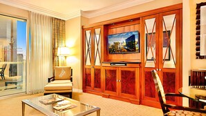 A hotel room with a large flat-screen TV mounted on a wooden cabinet, a comfortable armchair, a glass coffee table with magazines, and a view of the city through the window. - Amalz # 4, MGM Signature 2 Bedroom 3 Bathroom Balcony Suites ! (Las Vegas)