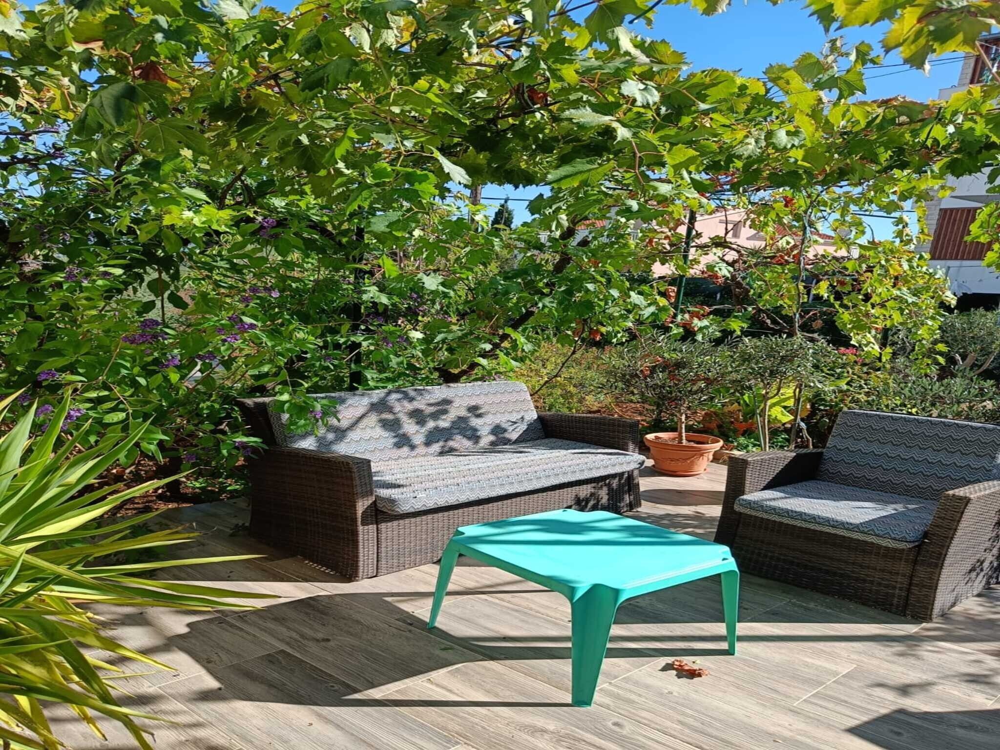An outdoor seating area with wicker furniture and a turquoise table, surrounded by lush greenery and a clear sky.