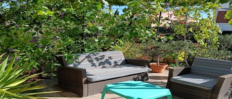 Uma área externa com assentos de vime e uma mesa turquesa, cercada por vegetação exuberante e céu claro.