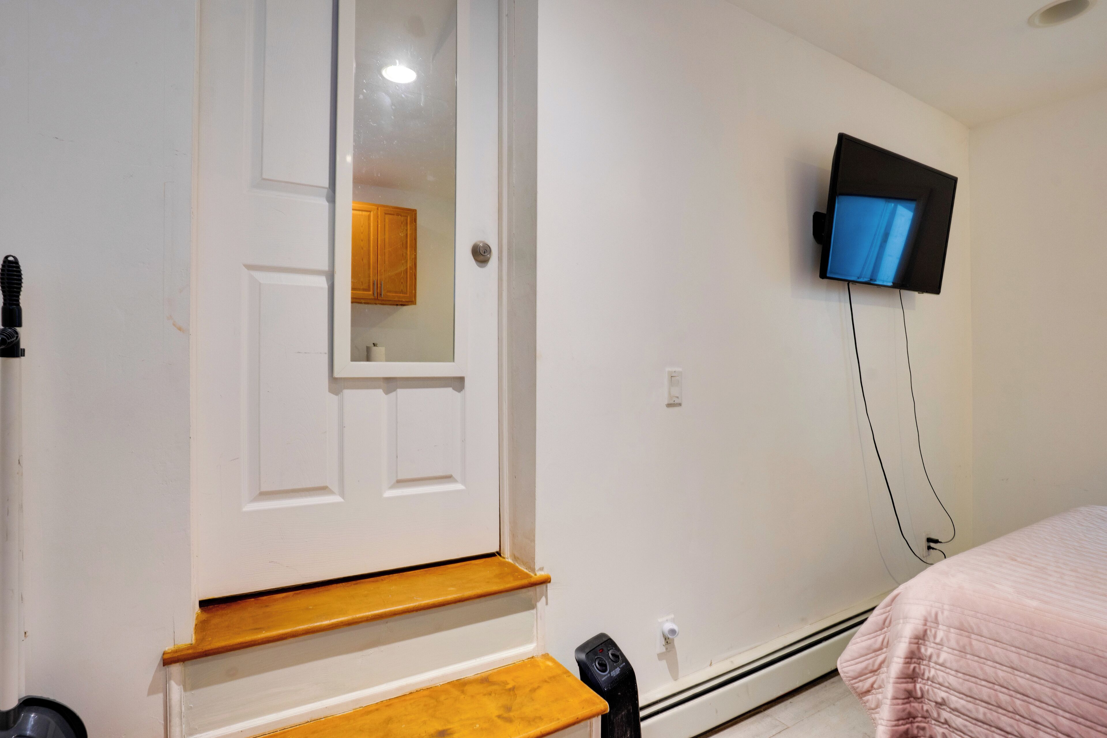 A room with a white door, wooden steps, a television mounted on the wall, and a bed with a pink blanket.