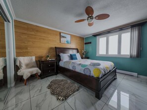 Quarto com cabeceira de madeira, cama com roupa de cama estampada, tapete de onça, mesa de cabeceira com um vaso e um ventilador de teto.