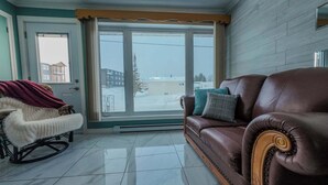 Ein Wohnzimmer mit einem braunen Ledersofa, einem Schaukelstuhl mit Felldecke und einem großen Fenster mit Blick auf eine verschneite Landschaft.