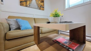 A modern living room with a sofa, a coffee table, a painting, a window, and a potted plant. - Logis near university, parking & Wi-Fi (Rouyn-Noranda)