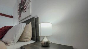 A neatly made bed with a red throw blanket, a beige pillow, and a white pillow. A modern table lamp with a white shade is on a dark wooden nightstand beside the bed. - Logis near university, parking & Wi-Fi (Rouyn-Noranda)