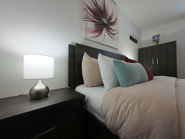 A neatly made bed with pillows, a bedside lamp, a dark wooden nightstand with drawers, and a large abstract painting on the wall. - Logis near university, parking & Wi-Fi (Rouyn-Noranda)
