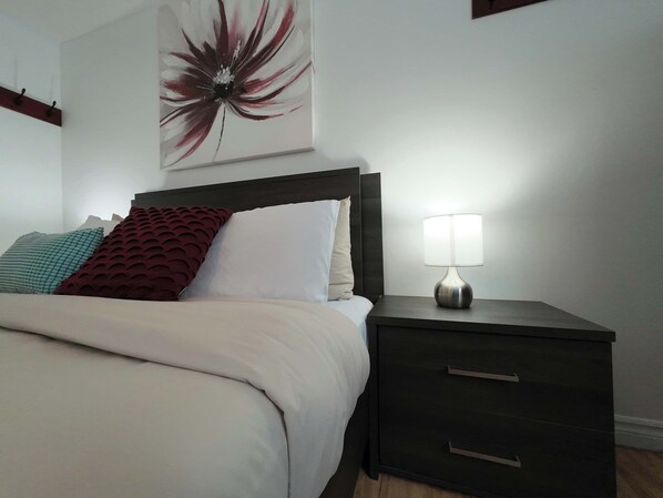 A neatly made bed with a white comforter and pillows, a dark wooden nightstand with a lamp, and a wall-mounted artwork. - Logis near university, parking & Wi-Fi (Rouyn-Noranda)