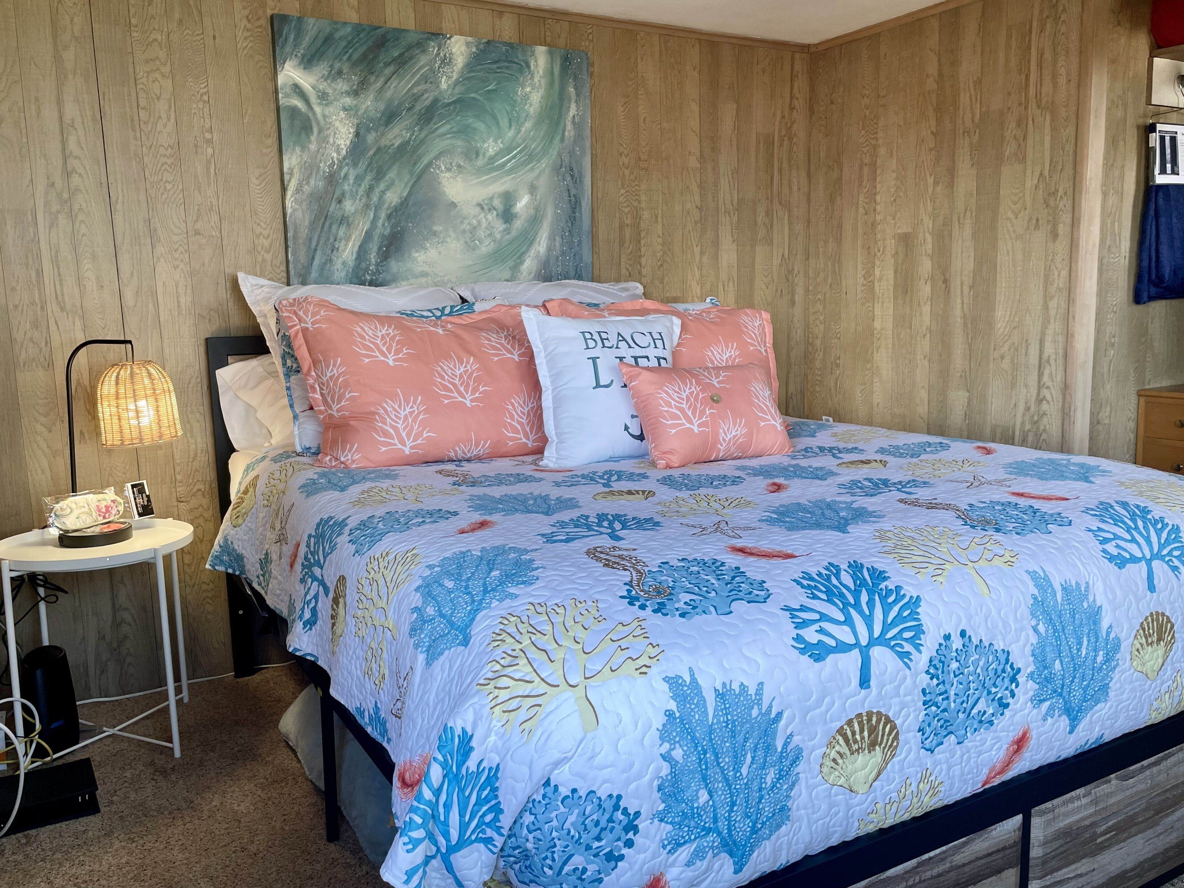A bedroom with a bed featuring a blue and white coral pattern bedspread, two pink pillows with tree designs, a white pillow with 'BEACH LIFE' text, and a wooden headboard.
